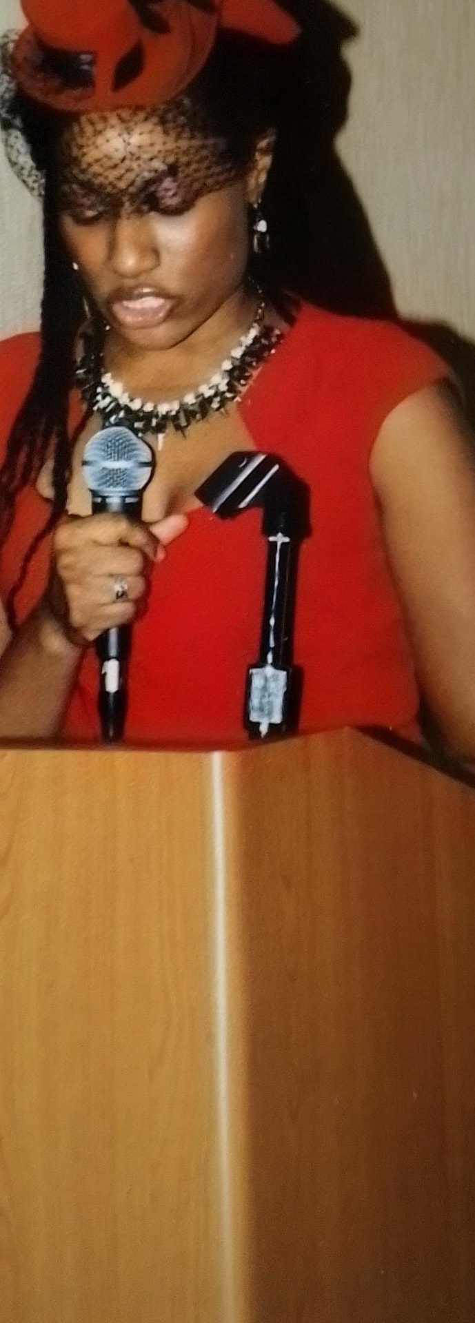 Sunni LaVasiette speaking at a podium in a red dress and hat